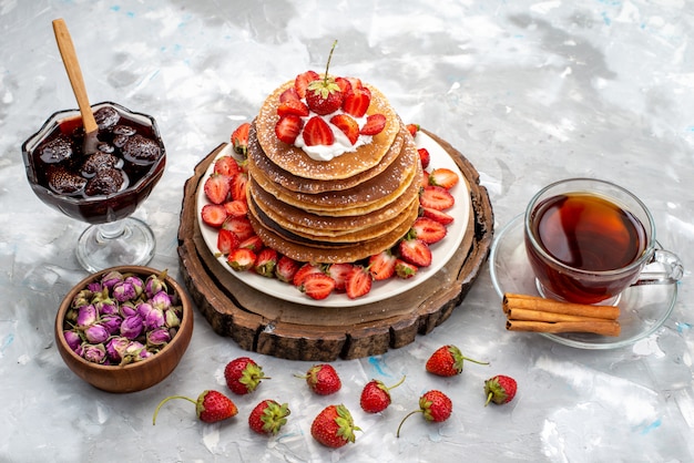 Una vista frontal deliciosos panqueques redondos con crema y té de fresas rojas en el pastel de escritorio de madera