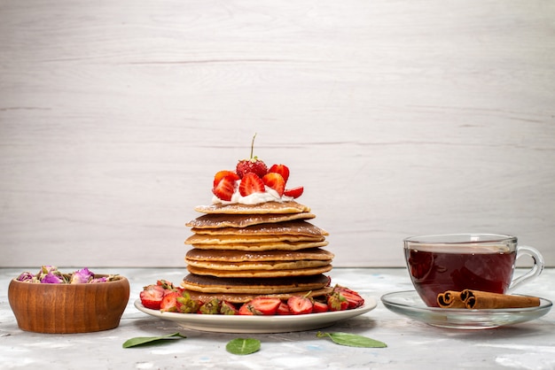 Una vista frontal deliciosos panqueques redondos con té crema y fresas rojas en el escritorio ligero pastel de frutas