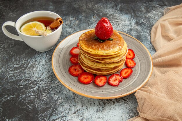 Vista frontal deliciosos panqueques con taza de té y frutas en la superficie de la luz de la torta de frutas dulces