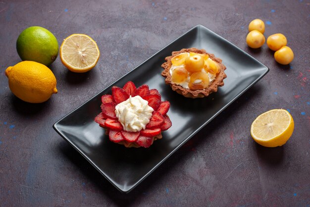 Vista frontal deliciosos pasteles cremosos dentro de la placa con limones frescos en la superficie oscura