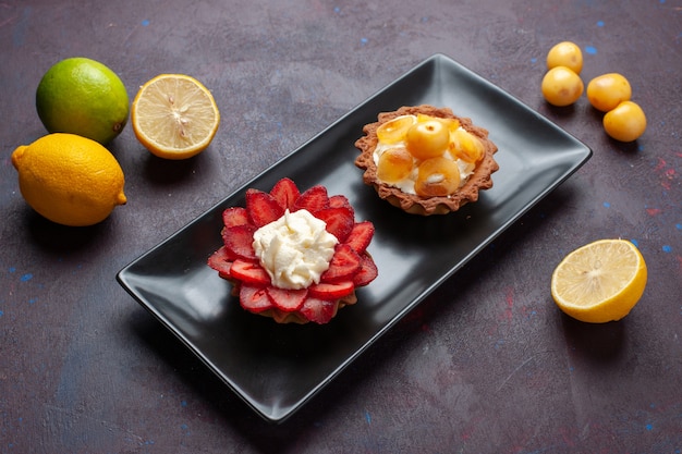 Vista frontal deliciosos pasteles cremosos dentro de la placa con limones frescos en la superficie oscura