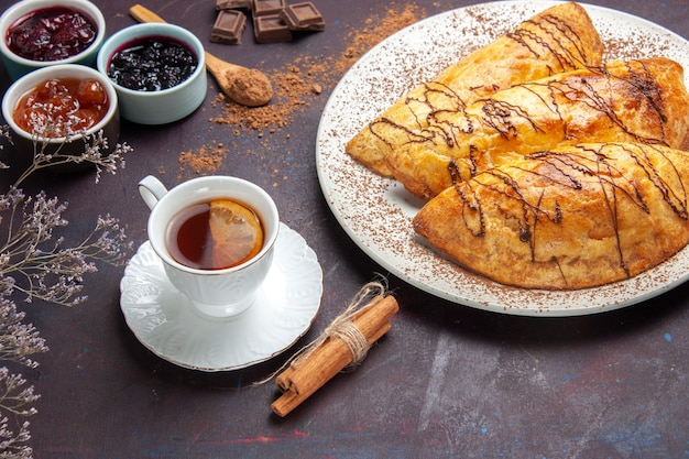 Vista frontal deliciosos pasteles horneados con taza de té y mermelada en el espacio oscuro