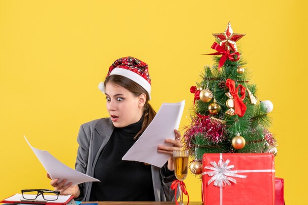 Vista frontal desconcertado niña con sombrero de Navidad sentado en la mesa mirando documentos árbol de Navidad y cóctel de regalos