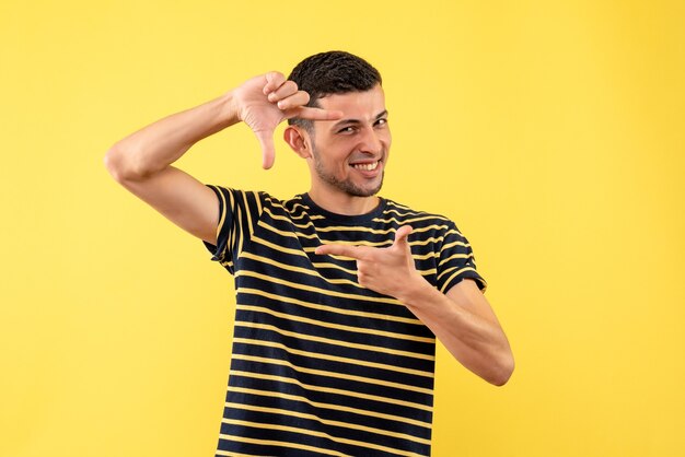 Vista frontal dichoso apuesto hombre en camiseta a rayas blanco y negro haciendo signo de cámara sobre fondo amarillo aislado