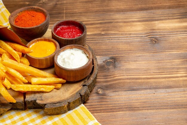 Vista frontal de diferentes condimentos con papas fritas en la comida de comida rápida de patata de mesa de madera marrón