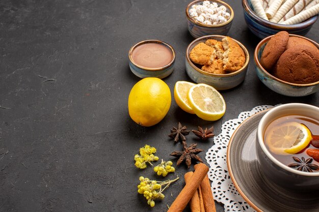 Vista frontal de diferentes galletas con una taza de té y rodajas de limón sobre fondo oscuro cookies galleta cítrica dulce fruta azúcar