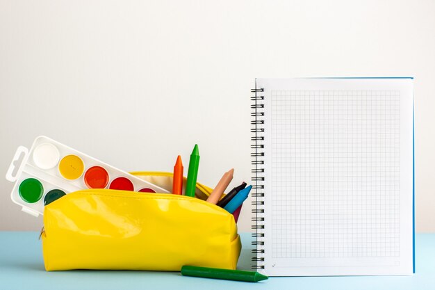 Vista frontal de diferentes lápices de colores dentro de la caja de lápiz amarillo con cuaderno sobre el escritorio azul