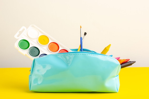 Vista frontal de diferentes lápices de colores y pinturas dentro de la caja de lápiz azul en el escritorio amarillo