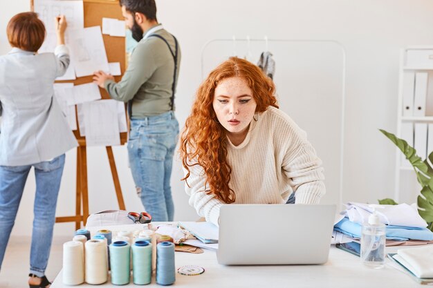 Vista frontal de la diseñadora de moda femenina que trabaja en el atelier con una computadora portátil y colegas