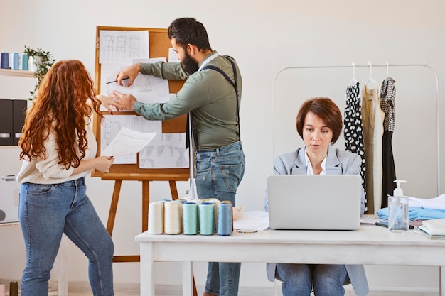 Vista frontal de la diseñadora de moda femenina que trabaja en el atelier con una computadora portátil y colegas