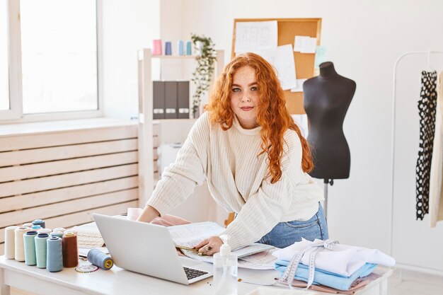 Vista frontal de la diseñadora de moda femenina que trabaja en el taller con un portátil