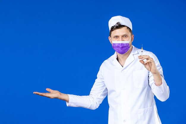 Vista frontal del doctor de sexo masculino sonriente en traje médico y máscara que sostiene la inyección en azul
