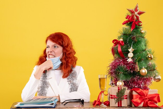 Vista frontal de la doctora alrededor del árbol de Navidad y presenta sentado en una máscara estéril