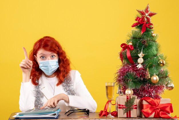 Vista frontal de la doctora alrededor del árbol de Navidad y presenta sentado en una máscara estéril