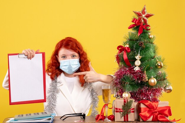 Vista frontal doctora alrededor del árbol de Navidad y presenta sentado en máscara