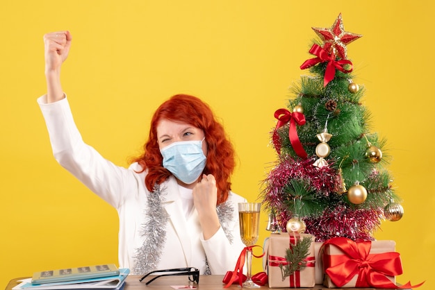 Foto gratuita vista frontal doctora alrededor del árbol de navidad y presenta sentado en máscara