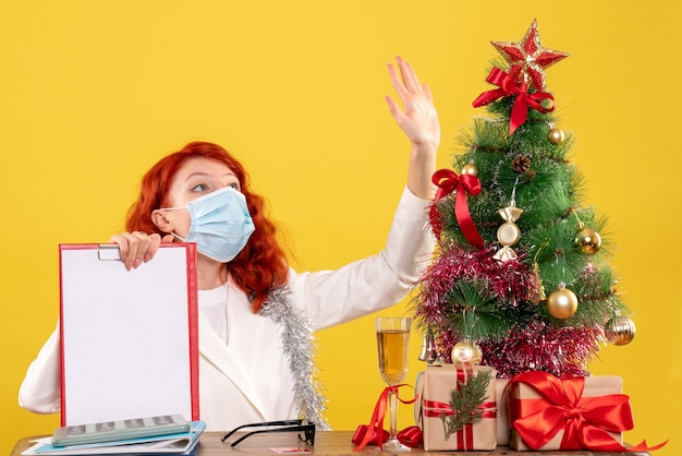 Vista frontal doctora alrededor del árbol de Navidad y presenta sentado en máscara