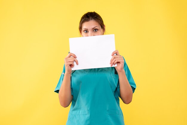 Vista frontal doctora en camisa médica con análisis de papel sobre fondo amarillo