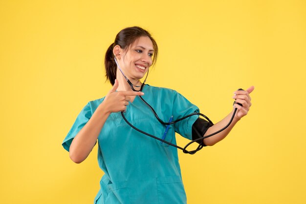 Vista frontal doctora en camisa médica comprobando su presión sobre fondo amarillo