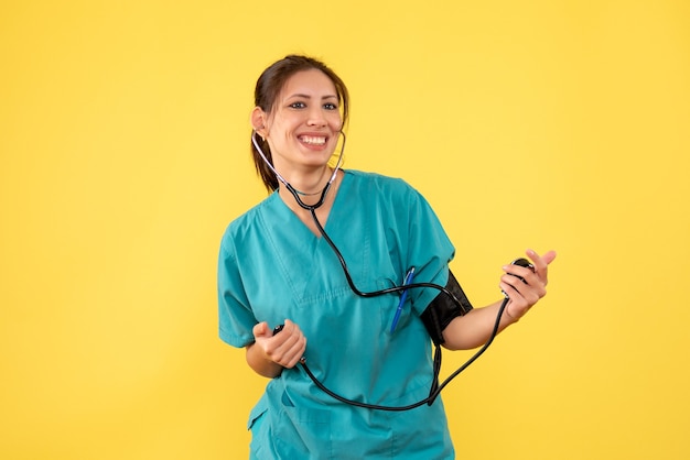 Vista frontal doctora en camisa médica comprobando su presión sobre fondo amarillo