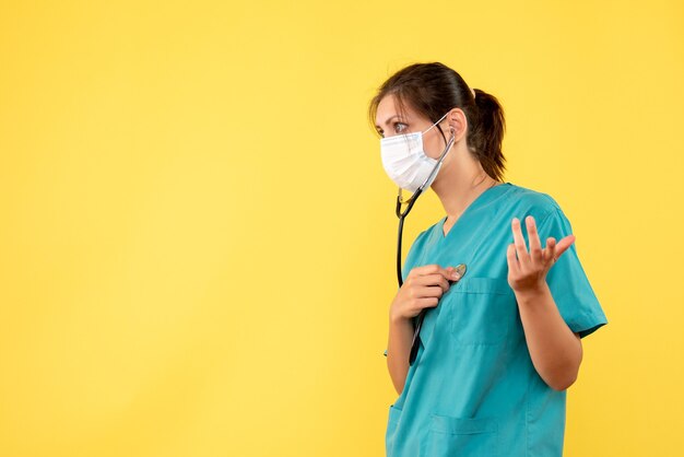 Vista frontal doctora en camisa médica y máscara con estetoscopio sobre fondo amarillo