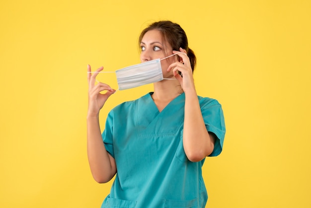 Vista frontal doctora en camisa médica quitándose la máscara sobre fondo amarillo