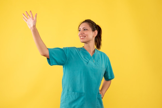 Vista frontal doctora en camisa médica saludando con una sonrisa sobre fondo amarillo