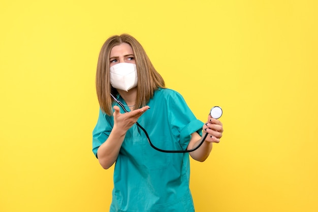 Vista frontal de la doctora con máscara y tonómetro en pared amarilla