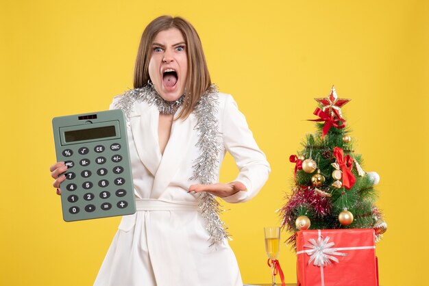 Vista frontal doctora de pie y con enojo sosteniendo la calculadora sobre fondo amarillo con árbol de navidad y cajas de regalo