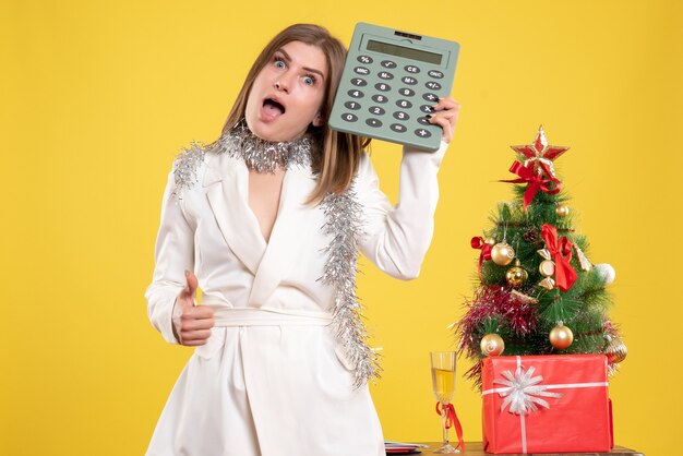 Vista frontal doctora de pie y sosteniendo la calculadora en amarillo con árbol de navidad y cajas de regalo