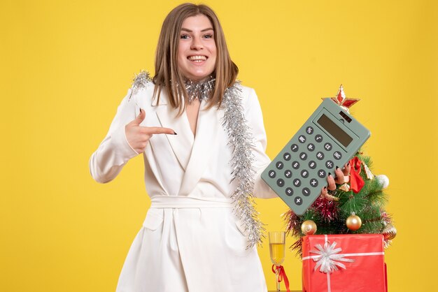Vista frontal doctora de pie y sosteniendo la calculadora en el escritorio amarillo con árbol de navidad y cajas de regalo