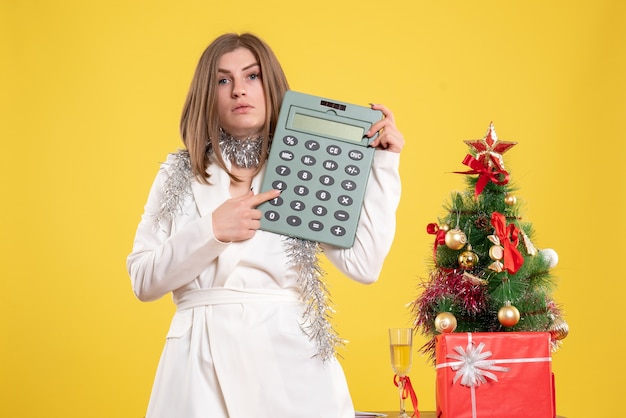 Vista frontal doctora de pie y sosteniendo la calculadora sobre fondo amarillo con árbol de navidad y cajas de regalo