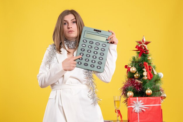 Vista frontal doctora de pie y sosteniendo la calculadora sobre fondo amarillo con árbol de navidad y cajas de regalo