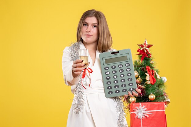 Vista frontal doctora de pie y sosteniendo la calculadora sobre fondo amarillo con árbol de navidad y cajas de regalo