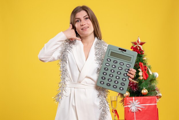 Vista frontal doctora de pie y sosteniendo la calculadora sobre fondo amarillo con árbol de navidad y cajas de regalo