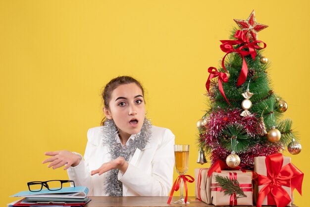 Vista frontal doctora sentada alrededor de regalos de Navidad y árbol confundido sobre fondo amarillo