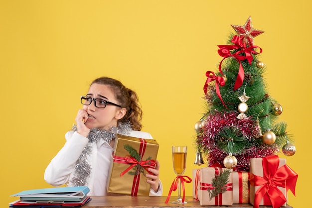 Vista frontal doctora sentada alrededor de regalos de Navidad y árbol nervioso sobre fondo amarillo