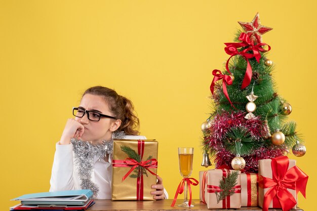 Vista frontal doctora sentada alrededor de regalos de Navidad y árbol pensando sobre fondo amarillo claro