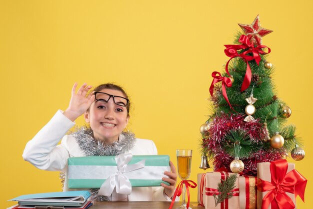 Vista frontal doctora sentada alrededor de regalos de Navidad y árbol con regalo sobre fondo amarillo