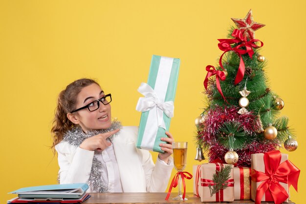 Vista frontal doctora sentada alrededor de regalos de Navidad y árbol con regalo sobre fondo amarillo