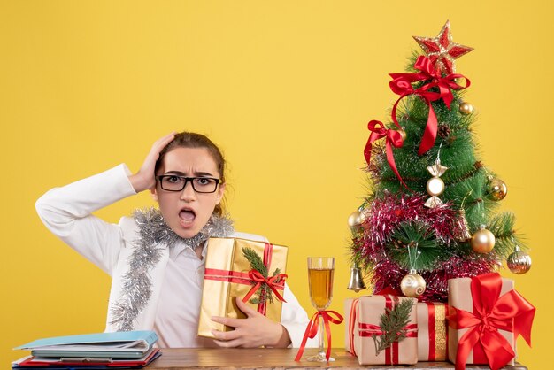 Vista frontal doctora sentada alrededor de regalos de Navidad y árbol con regalo sobre fondo amarillo