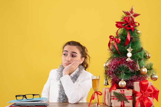 Vista frontal doctora sentada alrededor de regalos de Navidad y árbol triste sobre fondo amarillo