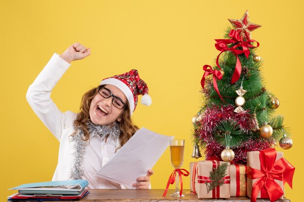 Vista frontal doctora sentada con árbol de regalos de navidad y sosteniendo documentos sobre fondo amarillo con árbol de navidad y cajas de regalo