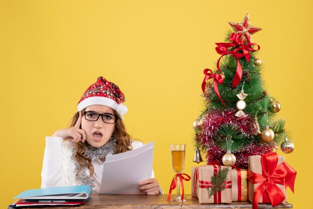 Vista frontal doctora sentada con árbol de regalos de Navidad y sosteniendo documentos sobre fondo amarillo