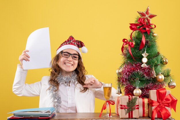 Vista frontal doctora sentada con árbol de regalos de Navidad y sosteniendo documentos sobre fondo amarillo