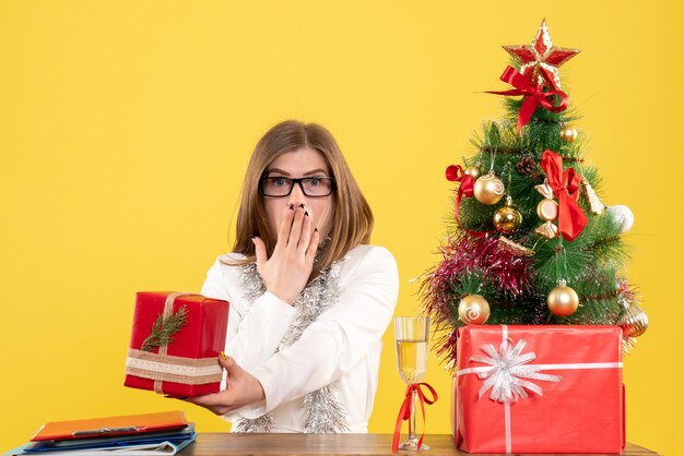 Vista frontal doctora sentada delante de la mesa con regalos y árbol sobre fondo amarillo con árbol de navidad y cajas de regalo