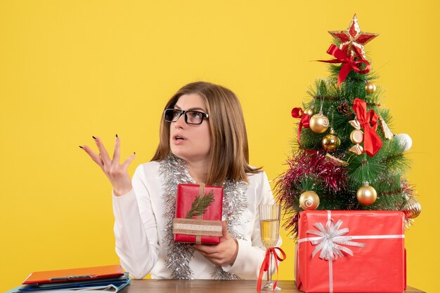 Vista frontal doctora sentada delante de la mesa con regalos y árbol sobre fondo amarillo con árbol de navidad y cajas de regalo