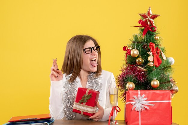 Vista frontal doctora sentada delante de la mesa con regalos y árbol sobre fondo amarillo con árbol de navidad y cajas de regalo