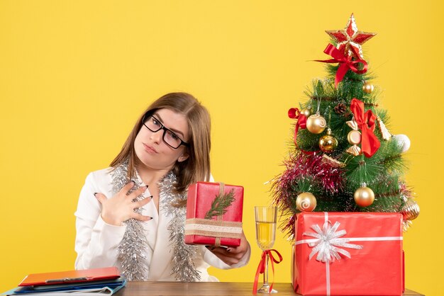 Vista frontal doctora sentada delante de la mesa con regalos y árbol sobre fondo amarillo con árbol de navidad y cajas de regalo