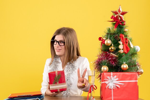 Vista frontal doctora sentada delante de la mesa con regalos y árbol sobre fondo amarillo con árbol de navidad y cajas de regalo